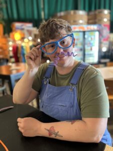 A white nonbinary person wearing overalls and a fake mustache smiles at the camera. This artist will lead a fun art workshop for LGBTQIA teens in St. Louis. 