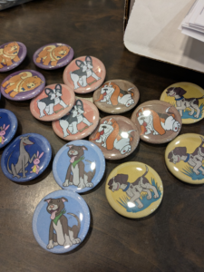 Buttons depicting dogs of various breeds are shown on a wood table. DIY button making is a fun art workshop for LGBTQIA+ middle and high schoolers in St. Louis Missouri.